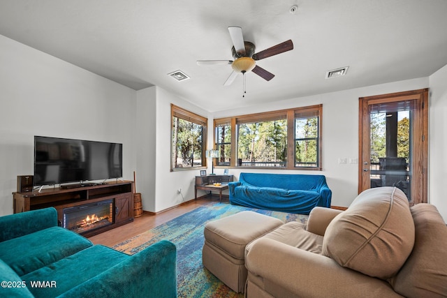 living area with a ceiling fan, a glass covered fireplace, wood finished floors, and visible vents