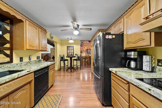 kitchen with light wood-type flooring, black appliances, a ceiling fan, under cabinet range hood, and light stone countertops