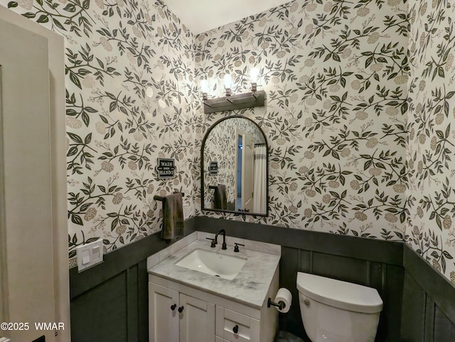 bathroom with wallpapered walls, toilet, vanity, and wainscoting