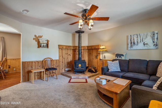 carpeted living area with arched walkways, ceiling fan, wood walls, wainscoting, and a wood stove