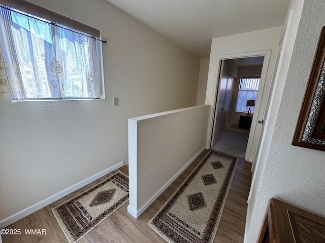 corridor with a textured wall, wood finished floors, an upstairs landing, and baseboards