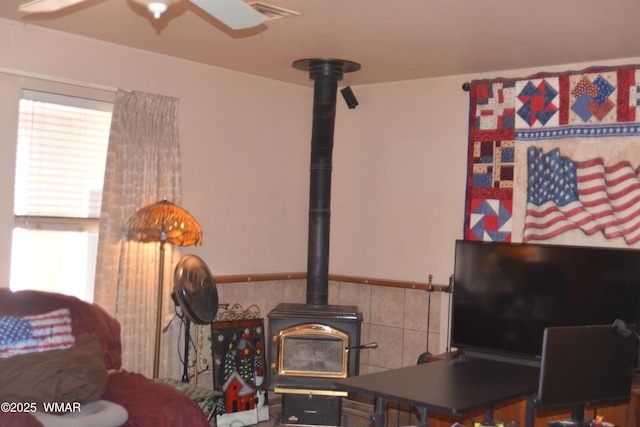 living area with a ceiling fan, visible vents, a wood stove, and a wainscoted wall