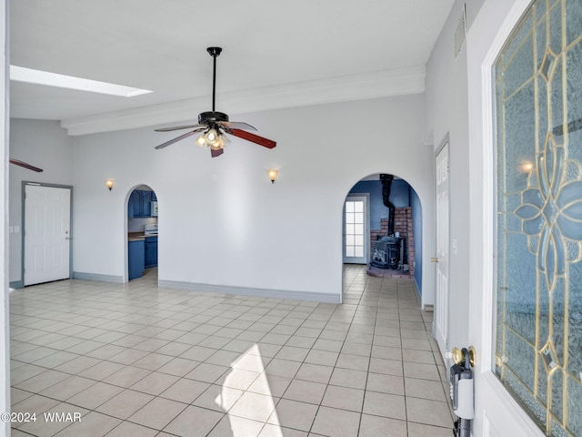 unfurnished room with vaulted ceiling with skylight, baseboards, ceiling fan, a wood stove, and light tile patterned flooring