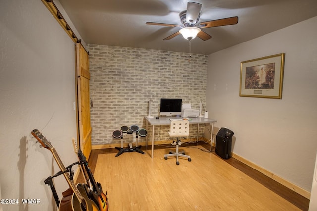 home office with ceiling fan, a barn door, brick wall, wood finished floors, and baseboards