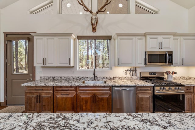 kitchen with appliances with stainless steel finishes, a sink, vaulted ceiling, light stone counters, and white cabinets