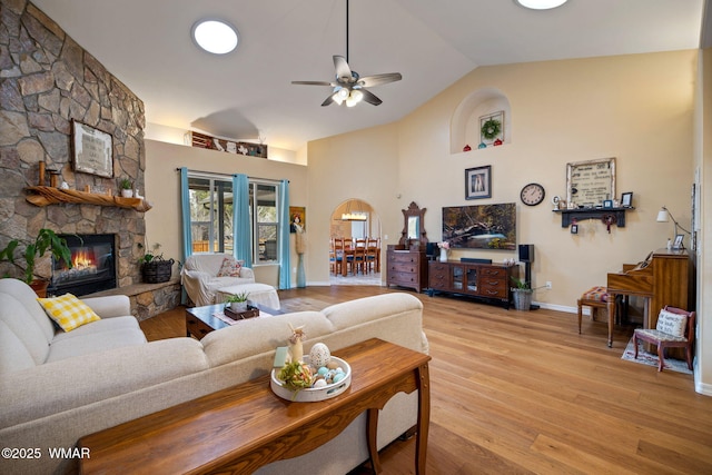 living area featuring light wood finished floors, baseboards, a stone fireplace, high vaulted ceiling, and a ceiling fan