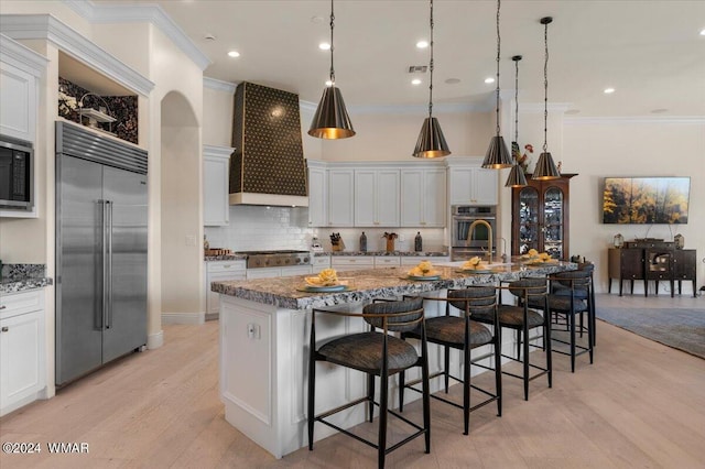 kitchen with a large island, hanging light fixtures, white cabinets, built in appliances, and premium range hood