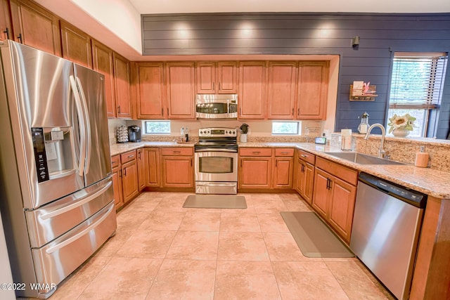 kitchen with light tile patterned floors, appliances with stainless steel finishes, brown cabinetry, a sink, and light stone countertops