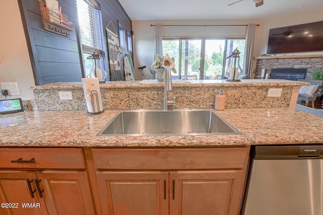 kitchen with brown cabinetry, a sink, a stone fireplace, light stone countertops, and dishwasher