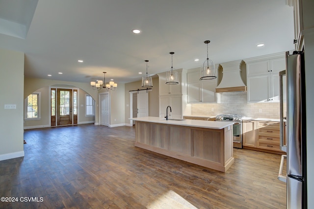 kitchen featuring appliances with stainless steel finishes, pendant lighting, custom range hood, hardwood / wood-style flooring, and a kitchen island with sink