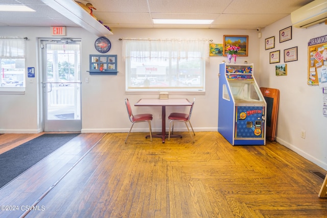 recreation room featuring an AC wall unit, a drop ceiling, a wealth of natural light, and hardwood / wood-style floors