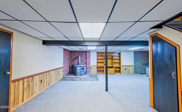 basement with a drop ceiling, carpet, wood walls, and a wood stove