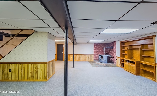 basement with a drop ceiling, wood walls, carpet flooring, and a wood stove