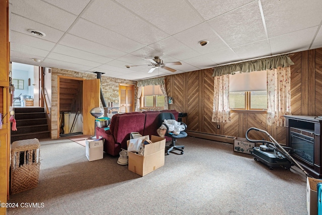 carpeted living room with a paneled ceiling, ceiling fan, wood walls, and a fireplace