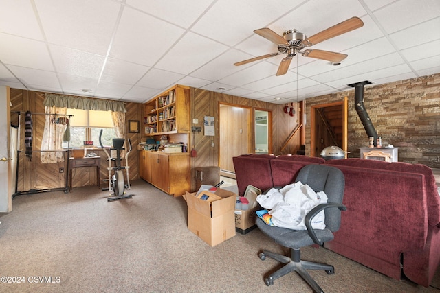 home office with carpet flooring, a wood stove, ceiling fan, a drop ceiling, and wooden walls