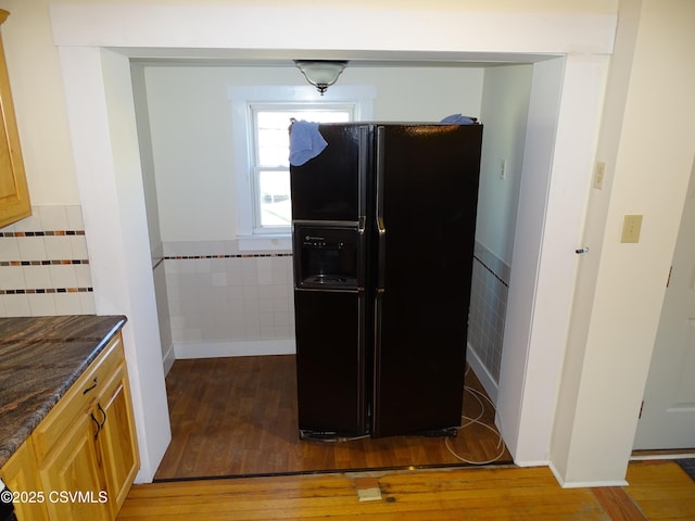 kitchen featuring black refrigerator with ice dispenser, light brown cabinetry, wood-type flooring, and tile walls