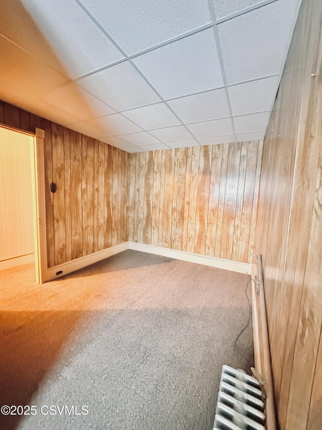 basement with carpet floors, a drop ceiling, radiator heating unit, and wooden walls