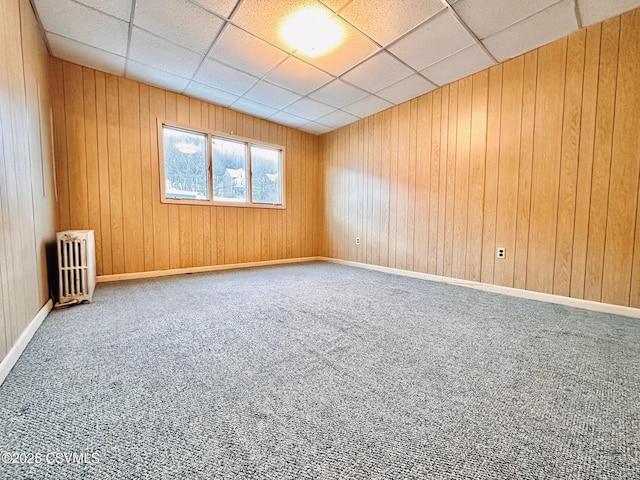 empty room with carpet floors, a drop ceiling, radiator, and wood walls