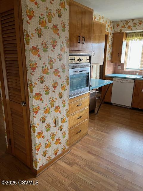 kitchen featuring stainless steel oven, dishwasher, light wood-style flooring, baseboards, and wallpapered walls