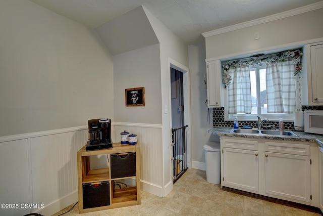 kitchen featuring wainscoting, light stone counters, ornamental molding, white cabinetry, and a sink