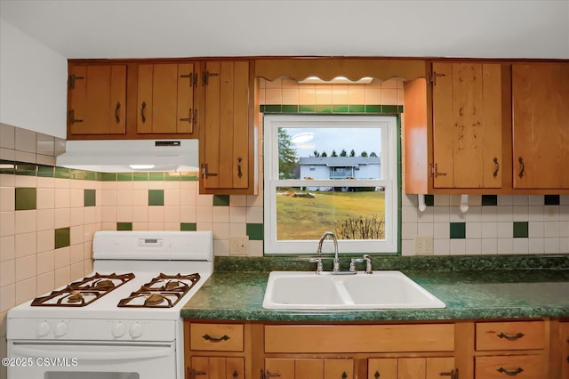 kitchen with dark countertops, decorative backsplash, a sink, under cabinet range hood, and white gas range oven