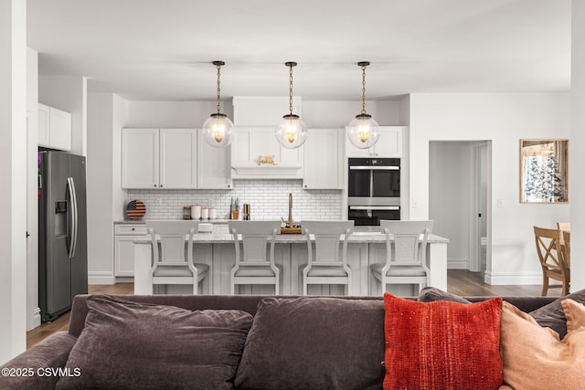 kitchen featuring a kitchen island with sink, stainless steel fridge, double oven, and decorative light fixtures