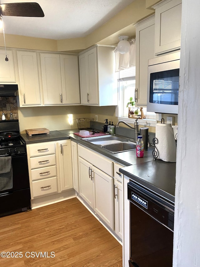 kitchen with under cabinet range hood, a sink, black appliances, light wood finished floors, and dark countertops