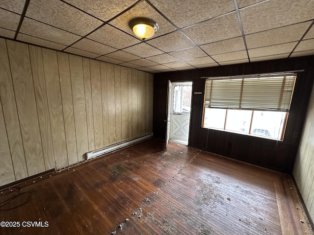 unfurnished room featuring dark wood-style floors, baseboard heating, a drop ceiling, and wooden walls