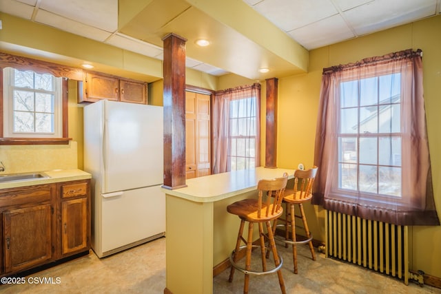 kitchen with radiator, freestanding refrigerator, a peninsula, light countertops, and a sink