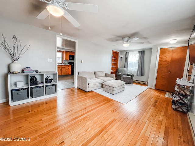 living room featuring baseboard heating, light wood-type flooring, a ceiling fan, and baseboards