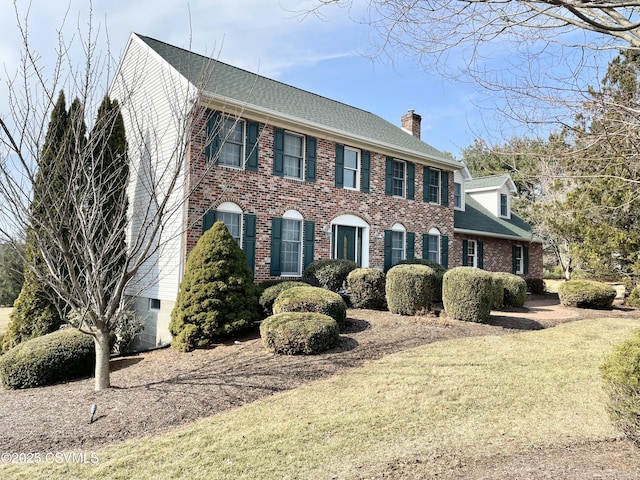 colonial-style house with a front yard, roof with shingles, a chimney, and brick siding