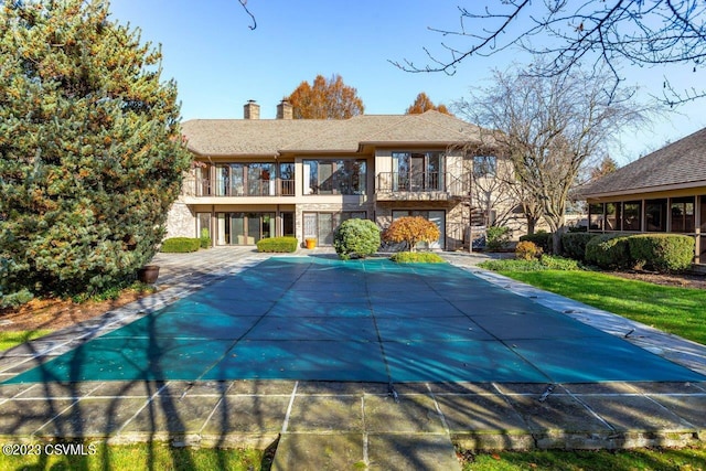 rear view of house featuring a balcony, stone siding, a covered pool, a chimney, and a patio area