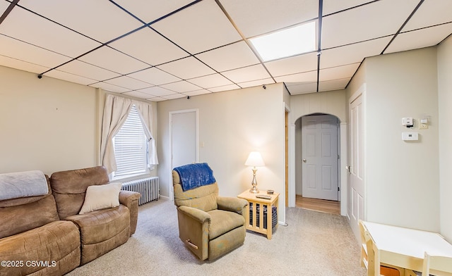 living area featuring a drop ceiling, arched walkways, radiator, and light carpet