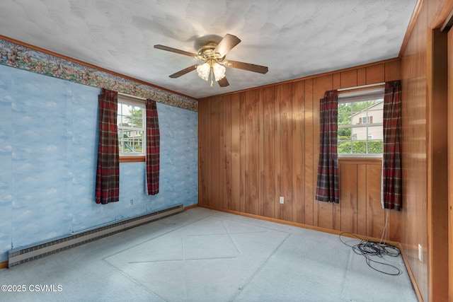 carpeted empty room featuring a baseboard radiator, plenty of natural light, ceiling fan, baseboards, and wooden walls