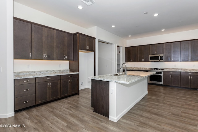 kitchen with appliances with stainless steel finishes, dark brown cabinets, sink, light stone counters, and a center island with sink