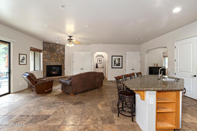 kitchen featuring a stone fireplace, dark stone countertops, an island with sink, sink, and a breakfast bar