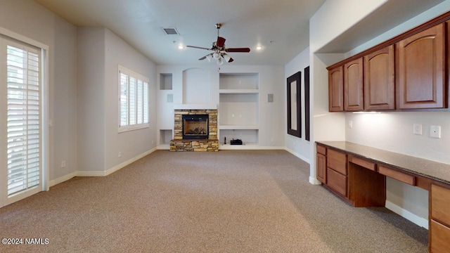 unfurnished living room with a fireplace, light carpet, built in desk, built in shelves, and ceiling fan