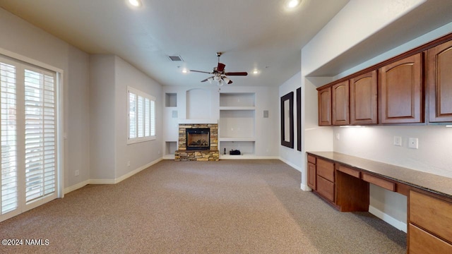 unfurnished living room featuring a fireplace, light colored carpet, ceiling fan, and built in desk