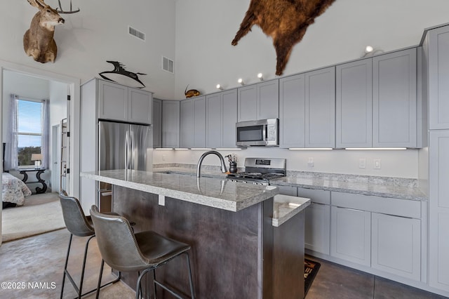 kitchen featuring high vaulted ceiling, stainless steel appliances, light stone countertops, and an island with sink