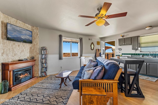 living area with visible vents, a ceiling fan, a glass covered fireplace, wainscoting, and light wood-type flooring