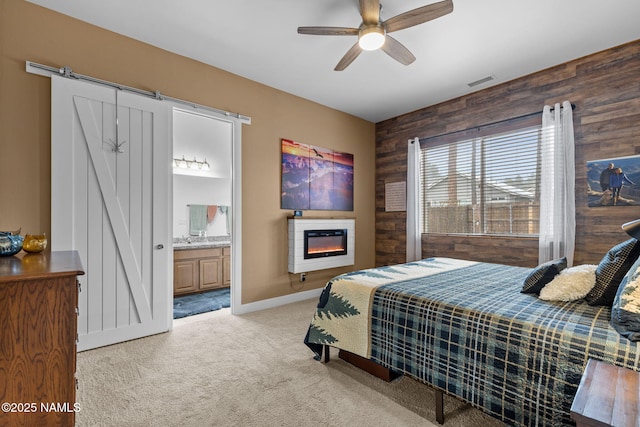 carpeted bedroom featuring a barn door, visible vents, a glass covered fireplace, ensuite bathroom, and wood walls