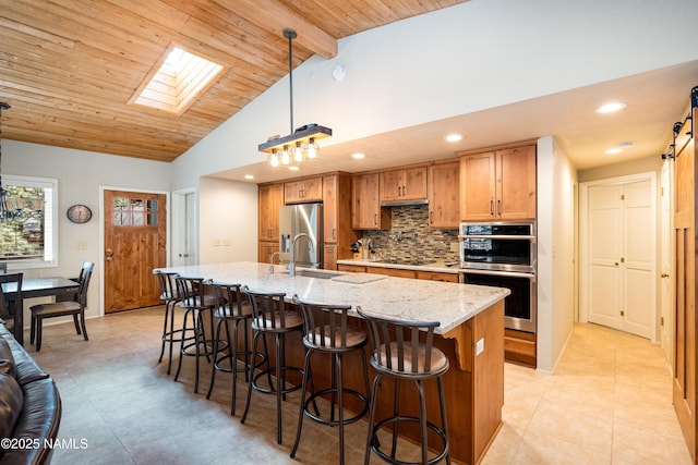 kitchen with an island with sink, sink, hanging light fixtures, light stone counters, and stainless steel appliances