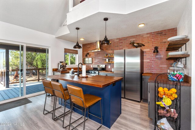 kitchen featuring wall chimney exhaust hood, wooden counters, decorative light fixtures, stainless steel appliances, and a breakfast bar