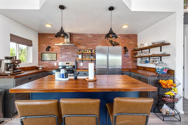 kitchen with appliances with stainless steel finishes, wall chimney range hood, hanging light fixtures, a breakfast bar, and butcher block countertops