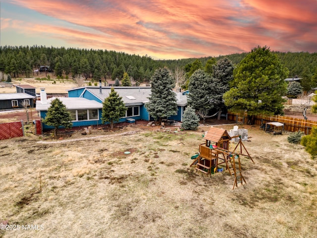 yard at dusk featuring a forest view, fence, and a playground