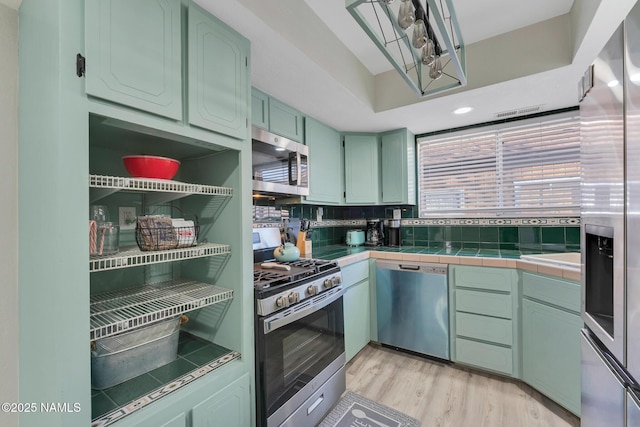 kitchen with stainless steel appliances, tile counters, green cabinetry, decorative backsplash, and light wood-type flooring