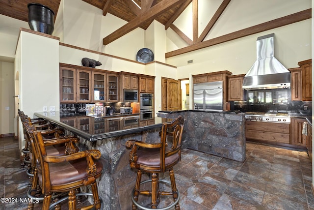 kitchen with wall chimney exhaust hood, built in appliances, tasteful backsplash, high vaulted ceiling, and a breakfast bar area