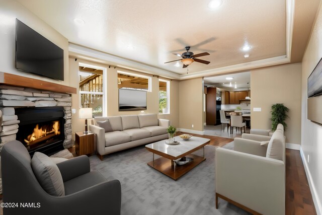 living room featuring a stone fireplace, crown molding, light hardwood / wood-style flooring, ceiling fan, and a tray ceiling