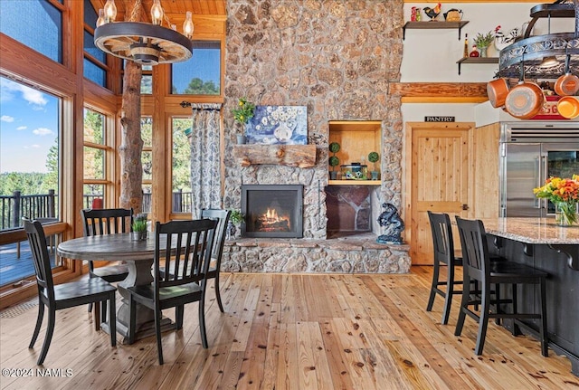 dining area with light wood-type flooring, a stone fireplace, a high ceiling, and an inviting chandelier