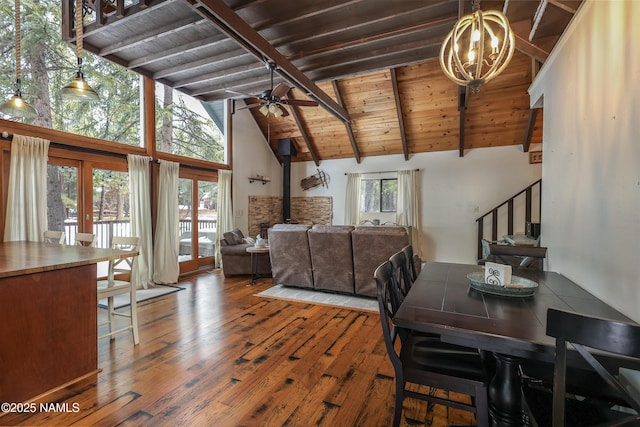 dining room featuring beamed ceiling, ceiling fan with notable chandelier, stairway, wood-type flooring, and a wood stove